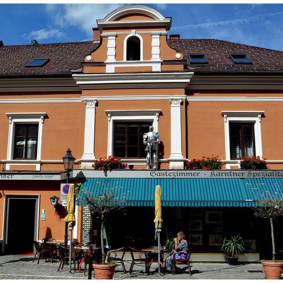 Ein Gebäude mit einem braunen Dach, weißen Verzierungen und einer Statue im zweiten Stock. Ein blaues Vordach bedeckt ein Café, in dem eine Frau auf einem Stuhl sitzt. Töpferpflanzen und Stühle stehen draußen.