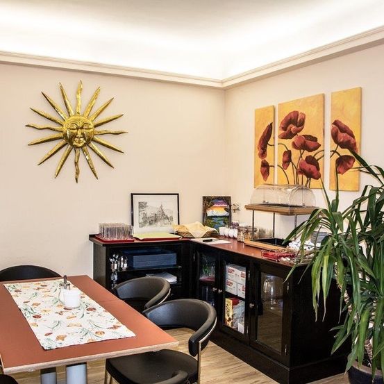 Ein Essbereich mit Tisch, Stühlen und einer Wand mit goldener Sonnendekoration. Es gibt auch einen Schrank mit verschiedenen Gegenständen und eine große Topfpflanze auf der rechten Seite.