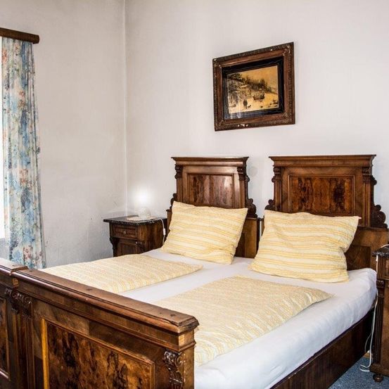 Ein Schlafzimmer mit zwei Holzbetten, die mit weißen Laken, gelben Kissen und einem blumengemusterten Vorhang geschmückt sind. Über den Betten hängt ein gerahmtes Bild an einer weißen Wand.