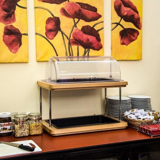 Ein Buffet-Display mit Glasdeckel steht auf einem Tisch. Es hat ein Holztablett, Stapel von Tellern und Gläser mit Lebensmitteln. Es gibt ein Messer auf einem Schneidebrett und einen Korb mit Servietten. Drei Gemälde roter Blumen hängen an der Wand.