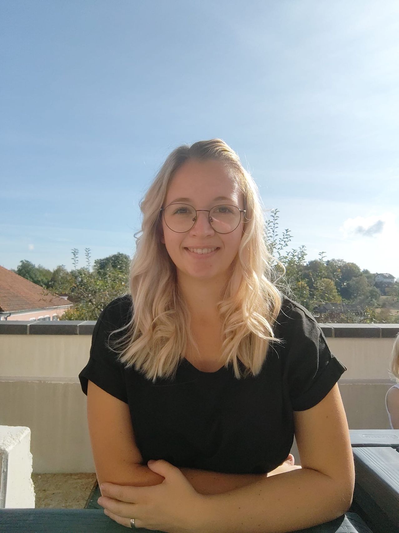 Eine Frau mit blonden Haaren und einer Brille lächelt in die Kamera. Sie steht draußen, mit Bäumen und einem Haus im Hintergrund.