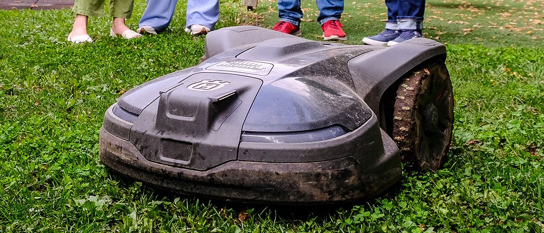 Vier Personen stehen in einem Park mit einem Roboter-Rasenmäher, in lässiger Kleidung. Der Rasenmäher steht auf dem Rasen, und hinter ihnen ist eine Schaukel.