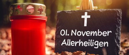 Eine rote Kerze steht neben einem Stein mit einem Kreuz und dem Text '01. November Allerheiligen'.