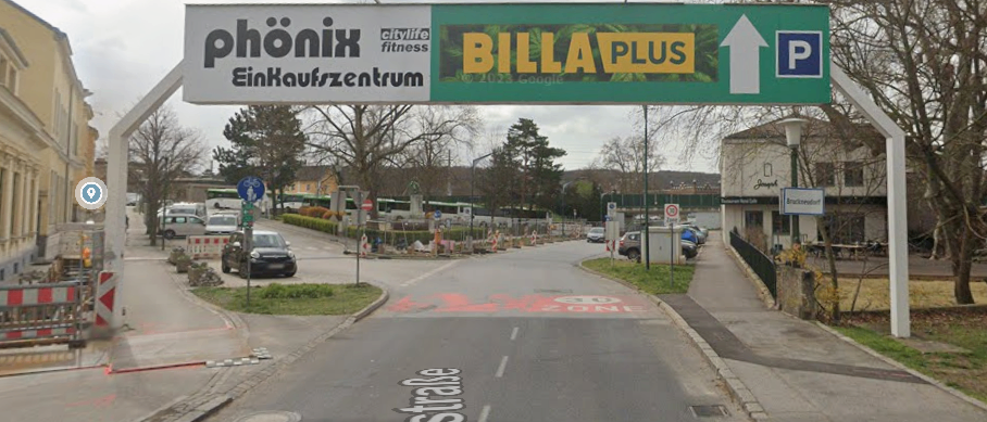 Eine Straßenszene mit einem großen Schild für das Phoenix Einkaufszentrum darüber, das auf Billa Plus und ein Parkzeichen hinweist. Straßenschilder, ein Zebrastreifen und geparkte Autos sind sichtbar, mit Bäumen und Gebäuden im Hintergrund.
