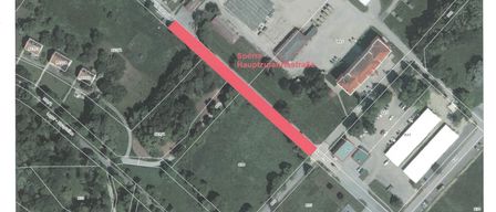 Eine Luftaufnahme eines Vorstadtgebiets zeigt eine Hauptstraße, die als Sperre Hauptzufahrtsstraße beschriftet ist. Die Karte enthält einen Maßstab und Entfernungsanzeigen. Es gibt Gebäude, Bäume und einen Parkplatz. Das Gebiet ist von Grün umgeben und umfasst andere Straßen und einen Parkplatz.