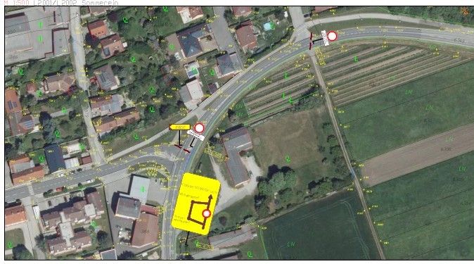 Aerial view of a town with several houses and a road. A yellow sign with a circular arrow points to the right. A red sign is also present.