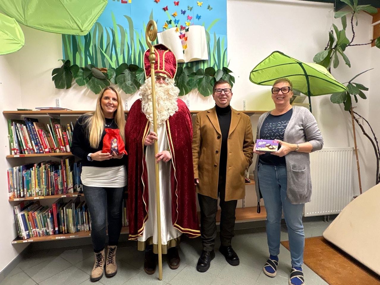 Drei Personen und ein Mann in der Kleidung des Heiligen Nikolaus stehen in einem Raum mit Bücherregalen und grünen Pflanzen. Die beiden Frauen halten Geschenke und der Mann einen Stab.