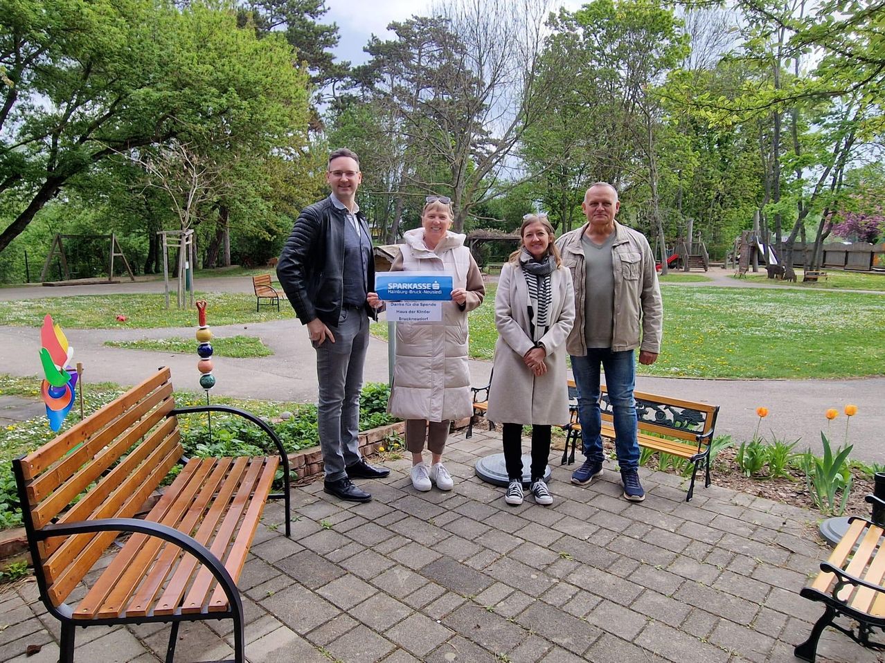 Drei Erwachsene stehen auf einem gepflasterten Weg in einem Park mit Bänken. Einer hält ein Zertifikat. Bäume und ein Spielplatz sind im Hintergrund.