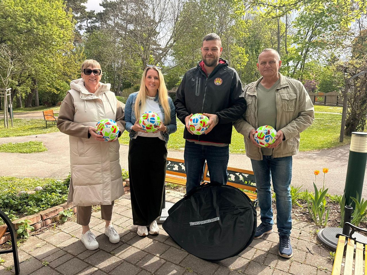 Vier Personen stehen in einem Park und halten mehrfarbige Fußballbälle. Ein Parkbank, Bäume und Pflanzen sind hinter ihnen.