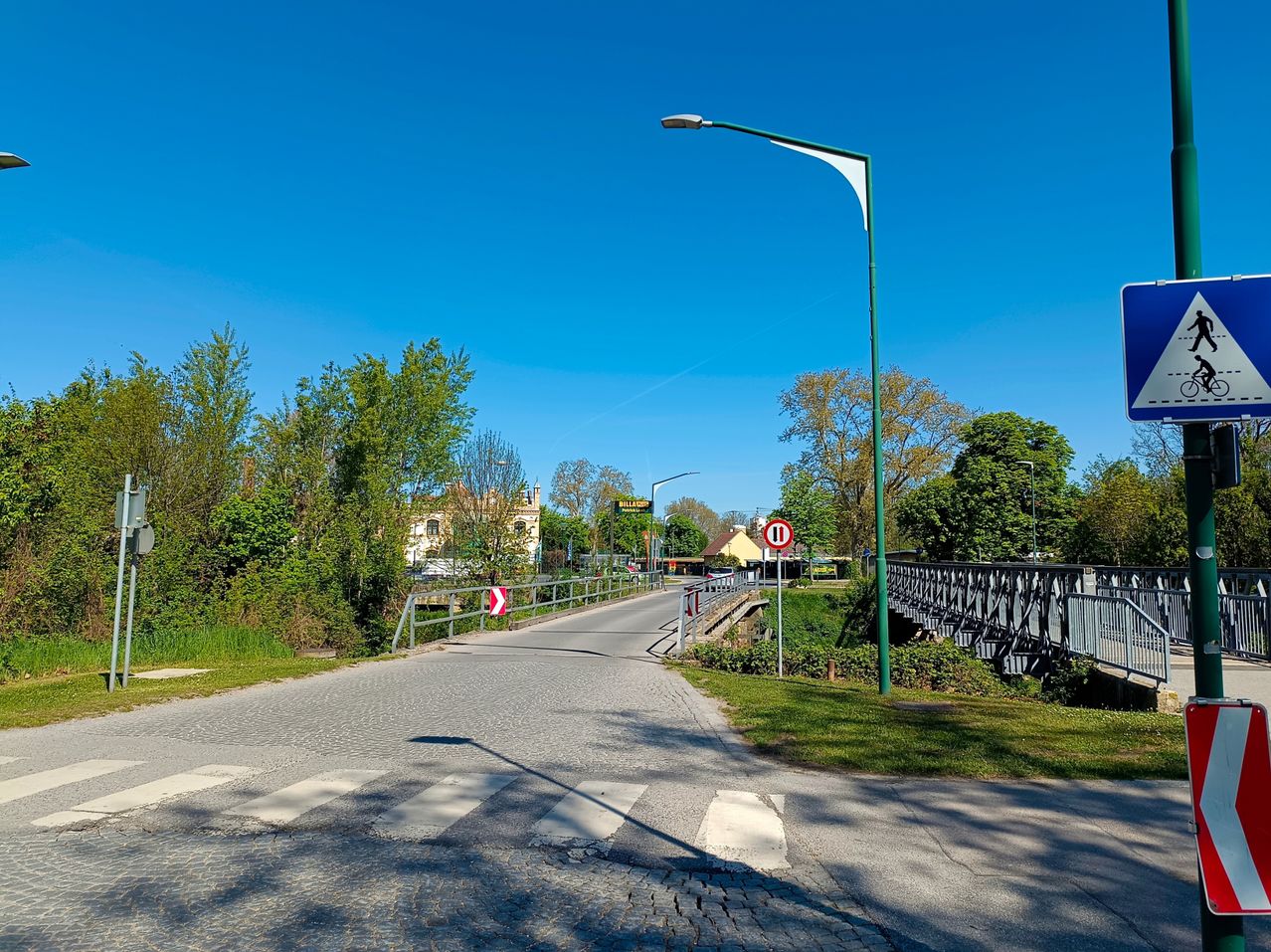 Eine leere Straße mit einem Zebrastreifen und Straßenlaternen. Eine Brücke und ein Gebäude sind in der Ferne. Bäume und Gras säumen die Seiten.