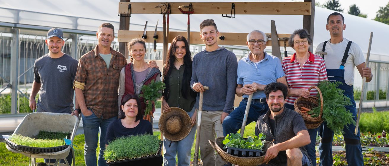 Eine Gruppe von Menschen steht draußen in einem Garten mit einem Pavillon im Hintergrund. Sie lächeln und posieren für ein Foto. Einige halten Pflanzen und Werkzeuge.