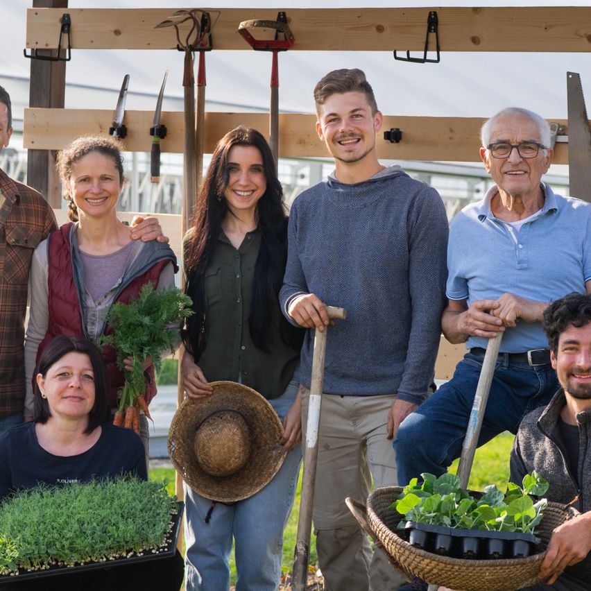 Eine Gruppe von Menschen steht draußen in einem Garten mit einem Pavillon im Hintergrund. Sie lächeln und posieren für ein Foto. Einige halten Pflanzen und Werkzeuge.