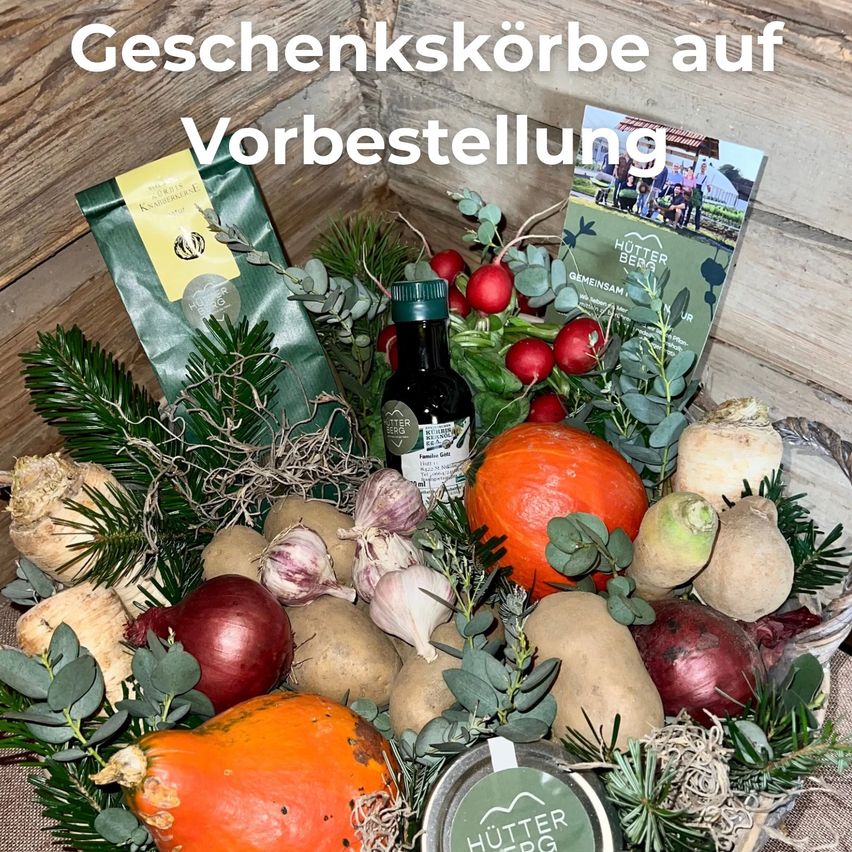 Auf einem hölzernen Hintergrund ist ein Geschenkkorb mit verschiedenen Gemüsesorten, Kräutern und einer Flasche zu sehen, alles mit grünen Blättern und Zweigen dekoriert. Ein Buch mit dem Titel 'Hütter Berg' ist ebenfalls enthalten.