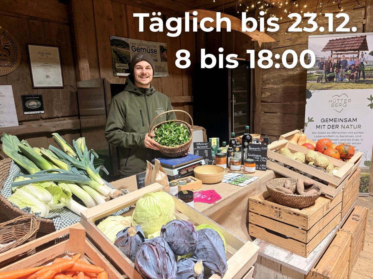 Ein lächelnder Mann steht an einem Gemüsestand und hält einen Korb mit Grünzeug. Verschiedenes Gemüse ist ausgestellt, und das Schild zeigt 'Täglich bis 23:00, 8 bis 18:00.' Die Umgebung ist im Freien mit Holzwänden.