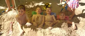 Vier Jungen liegen in einem Sandkasten, zwei tragen Mützen. Ein Mädchen in einem rosa Kleid steht hinter ihnen. Ein Junge mit einer grünen Mütze hält eine Schaufel und eine Frau in Shorts steht unter einem Regenschirm.