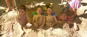 Four boys lie in a sandpit, two wearing hats. A girl in a pink dress stands behind them. A boy with a green hat holds a shovel, and a woman in shorts stands under an umbrella.