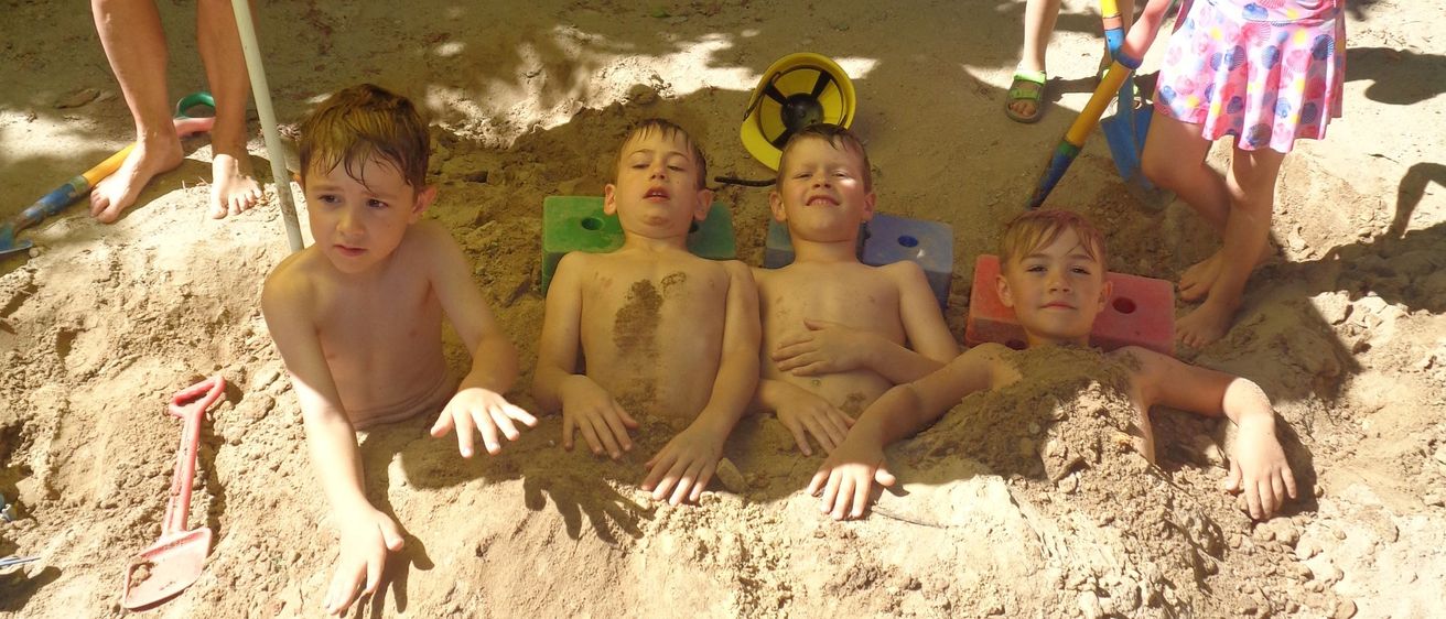 Vier Jungen liegen in einem Sandkasten, zwei tragen Mützen. Ein Mädchen in einem rosa Kleid steht hinter ihnen. Ein Junge mit einer grünen Mütze hält eine Schaufel und eine Frau in Shorts steht unter einem Regenschirm.