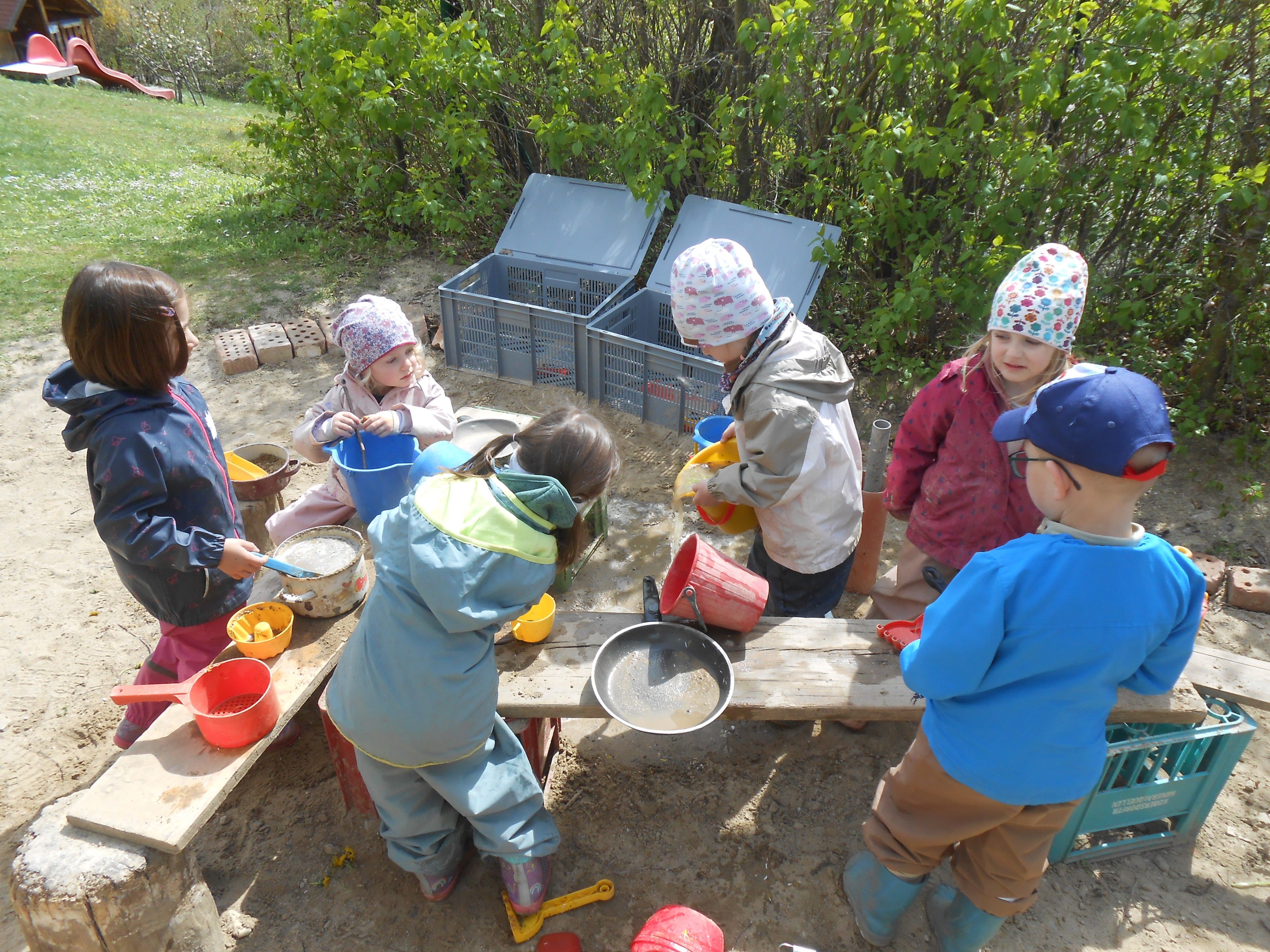 Kinder spielen draußen mit Sand und Wasser. Sie haben Eimer, Pfannen und Spielzeug. Ein Kind schaut zu. Hinter ihnen befinden sich Lagerboxen und Pflanzen.