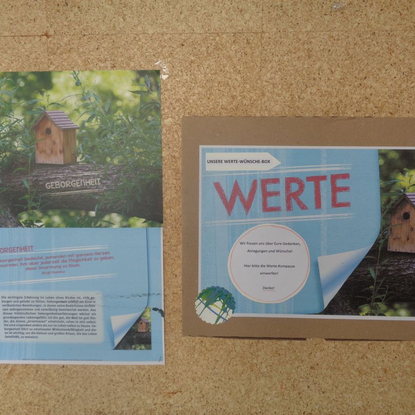 Ein Nahaufnahme eines blauen Plakats mit dem Wort 'Werte' in großen roten Buchstaben. Darunter befindet sich ein kleineres Plakat mit einem Vogelhäuschen-Bild und dem Text 'Geborgenheit'. Beide Plakate sind auf einer braunen Korkplatte.