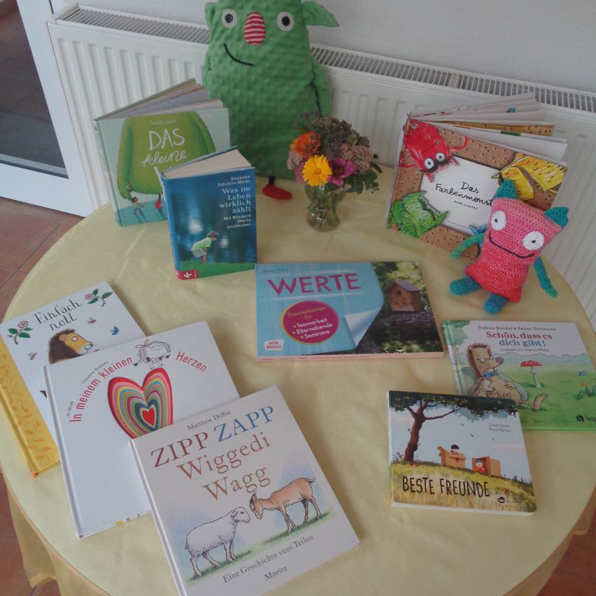 Ein Tisch mit mehreren Kinderbüchern, einem Blumenvase und zwei Plüschtieren. Die Bücher haben farbenfrohe Illustrationen und Titel. Der Tisch ist mit einer gelben Tischdecke bedeckt.