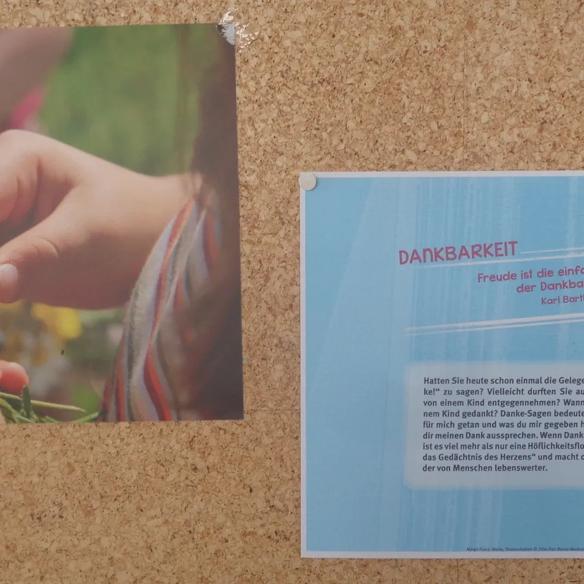 Die Hände eines Kindes, die einen Strauß Gänseblümchen halten, auf einer Pinnwand mit einem blauen Plakat mit Text auf Deutsch