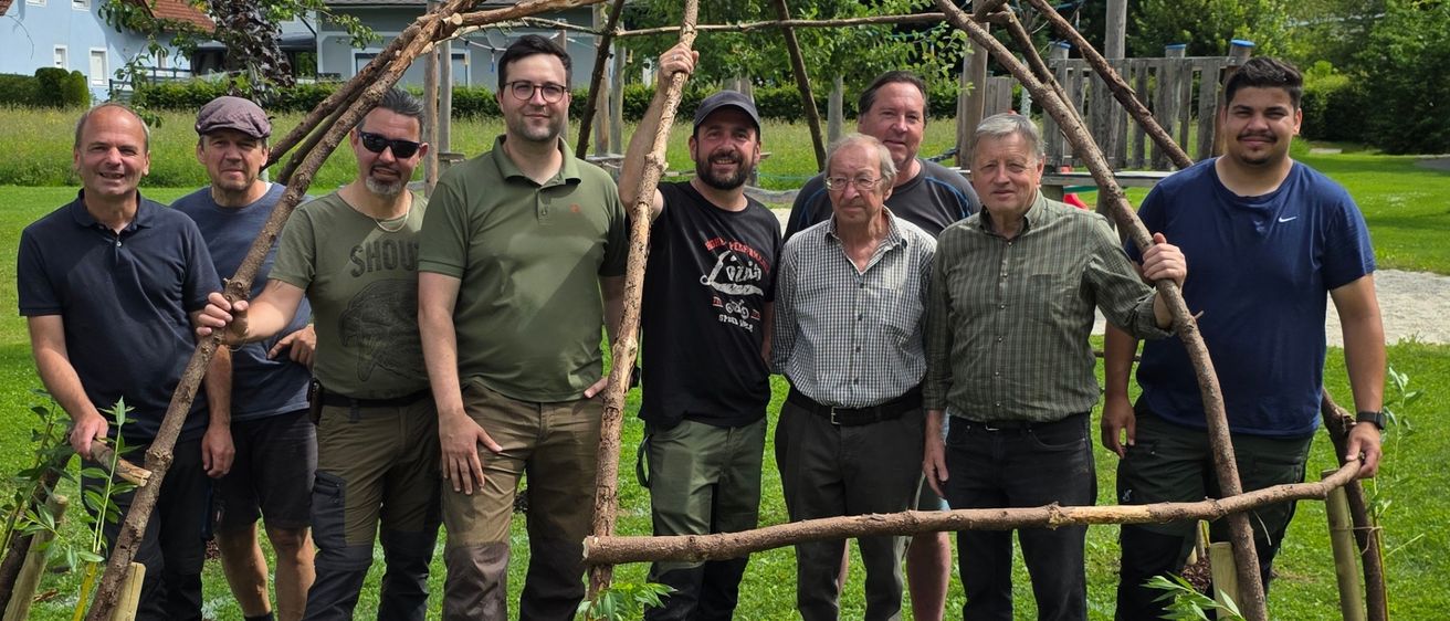 Eine Gruppe von Menschen steht um eine kleine Baumstruktur auf einem Rasenfeld. Sie scheinen für ein Foto zu lächeln.