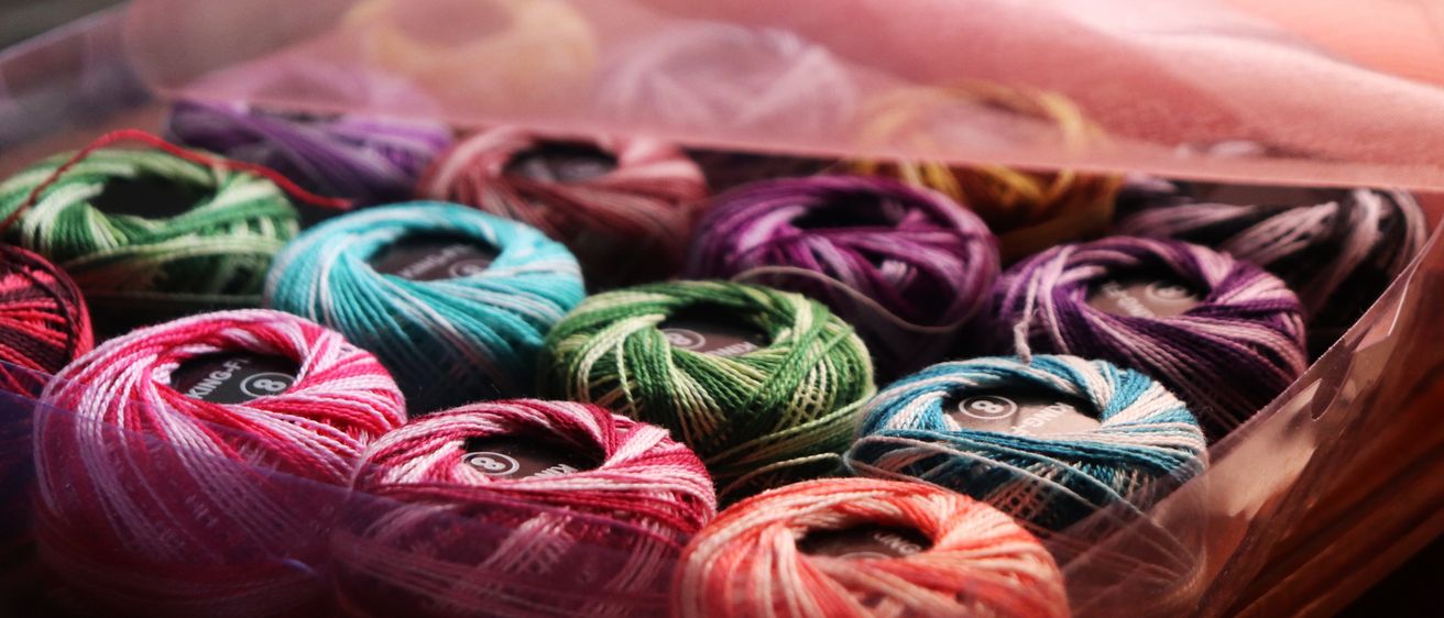 Ein Plastikbehälter mit Wollknäueln in verschiedenen Farben, einige mit Etiketten, liegt auf einem rosa Stoff.