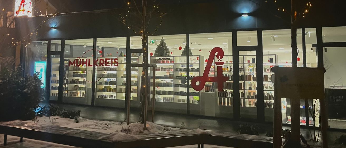 Ein Geschäft mit einem roten A-Logo ist nachts zu sehen. Die Schaufensterfront ist mit festlichen Lichtern und Weihnachtsdekorationen geschmückt. Schnee bedeckt den Boden und die nahegelegenen Bänke.