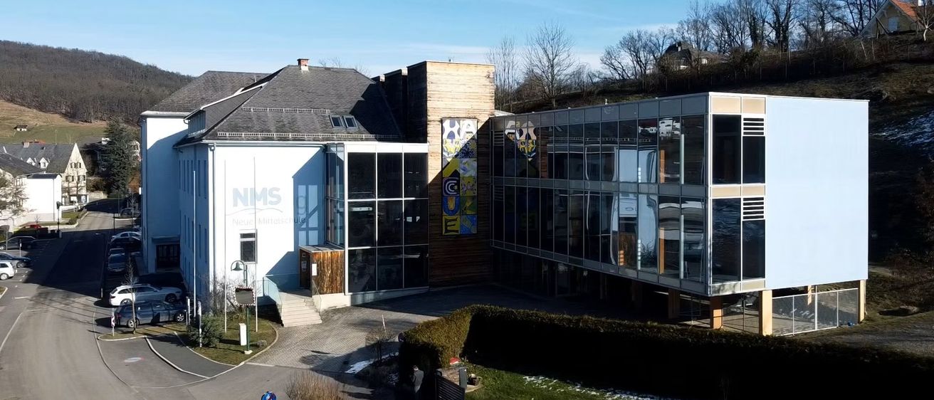 Ein modernes Gebäude mit Glaswänden und einem farbenfrohen Wandbild. Ein Weg führt zum Eingang, mit einem Schild und Sträuchern in der Nähe. Die Szene spielt sich unter einem klaren blauen Himmel ab.