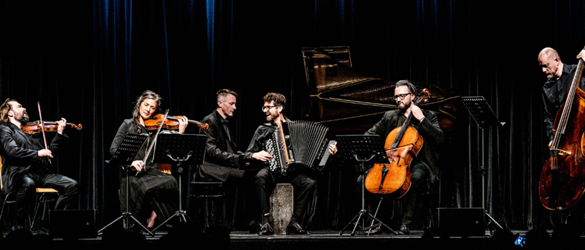 Eine kleine Musikergruppe spielt auf der Bühne. Die Geigerin und die Cellistin sind Frauen. Der Mann spielt Akkordeon. Dahinter steht ein Klavier.