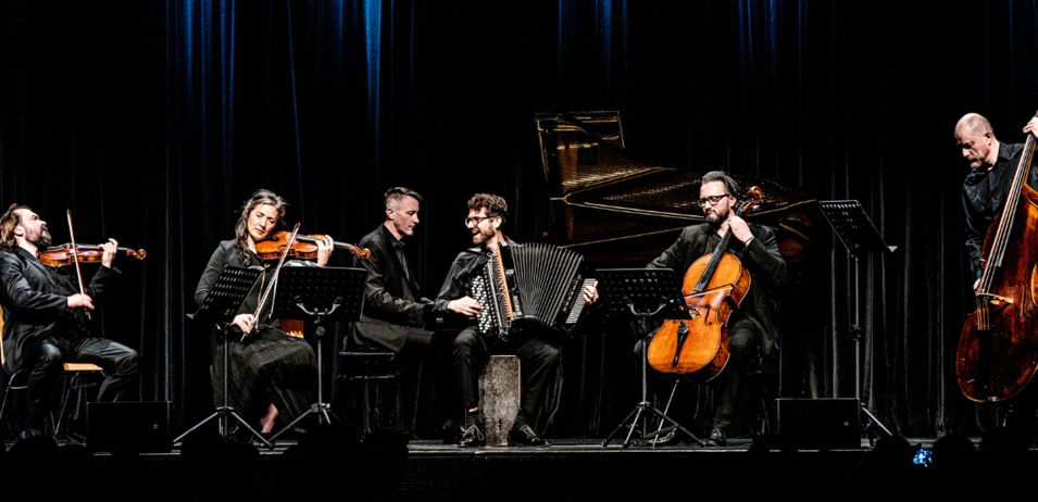 Eine kleine Musikergruppe spielt auf der Bühne. Die Geigerin und die Cellistin sind Frauen. Der Mann spielt Akkordeon. Dahinter steht ein Klavier.