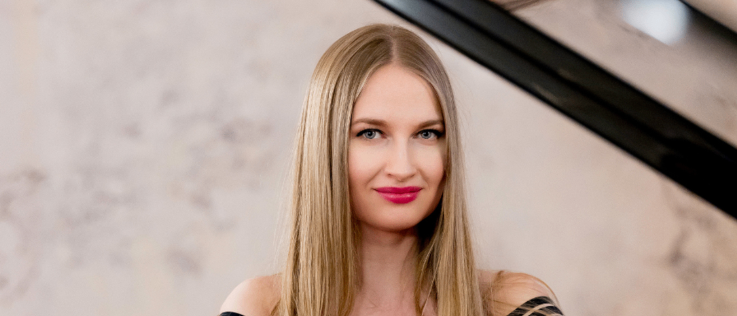 Eine Frau mit blonden Haaren sitzt am Klavier und lächelt. Sie trägt ein schwarzes ärmelloses Oberteil mit weißen Streifen. Das Klavier hat einen goldenen Rand und reflektiert ihr Bild.