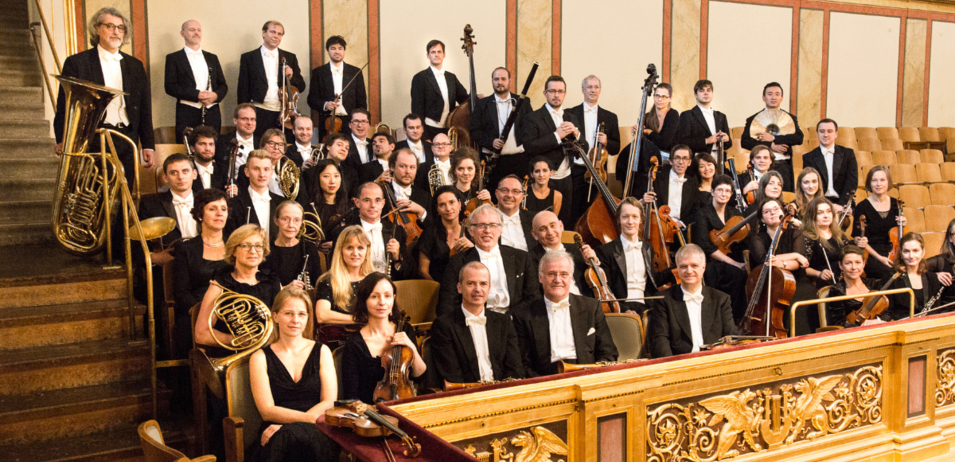 Ein großes Orchester versammelt sich in einem Konzertsaal, mit Musikern, die verschiedene Instrumente halten. Sie sind in formeller Kleidung gekleidet.