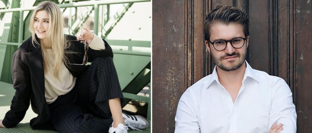 Eine Frau mit blondem Haar und einem schwarzen Outfit sitzt auf einer Brücke. Ein Mann mit Brille und weißem Hemd steht vor einer braunen Wand.