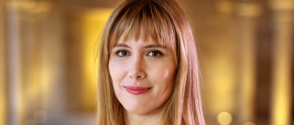 Eine Frau mit blonden Haaren, lächelnd und trägt ein schwarzes und weißes ärmelloses Oberteil mit einer Halskette. Der Hintergrund ist unscharf mit warmem gelbem Licht.