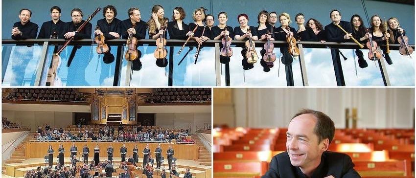 Eine Gruppe Musiker mit verschiedenen Instrumenten posiert auf einem Balkon über einem Konzertsaal. Unten spielt ein Orchester im Konzertsaal. Ein Mann lächelt in einem Auditorium.