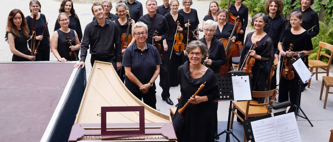 Eine Gruppe von Musikern steht draußen und lächelt für ein Foto. Geigen, Celli und ein Cembalo sind sichtbar. Der Hintergrund zeigt einen Brunnen und Grünpflanzen.