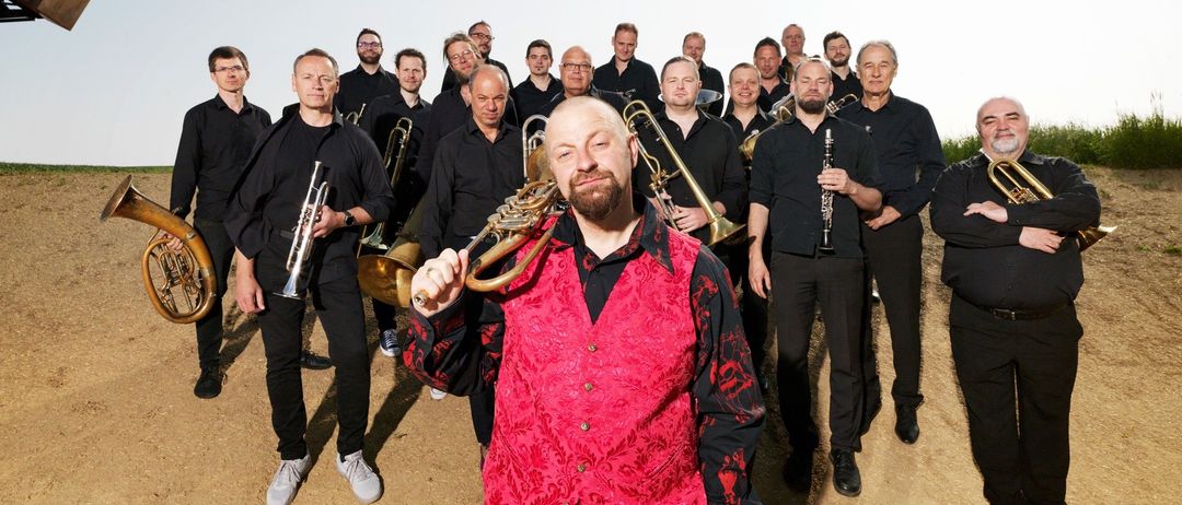 Eine Gruppe von Musikern posiert für ein Foto im Freien. Sie halten verschiedene Instrumente wie Trompeten und Posaunen.