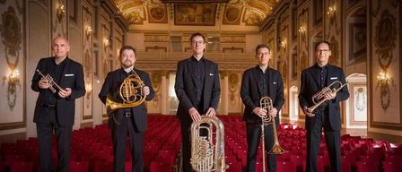 Fünf Männer stehen in einem großen Saal und halten Blechblasinstrumente. Der Saal hat eine hohe Decke mit kunstvollen Fresken. Rote Sitze füllen den Raum hinter ihnen.