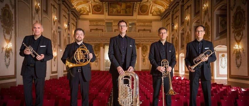Fünf Männer stehen in einem großen Saal und halten Blechblasinstrumente. Der Saal hat eine hohe Decke mit kunstvollen Fresken. Rote Sitze füllen den Raum hinter ihnen.