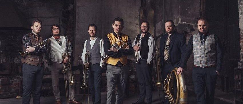 Eine Gruppe Musiker in formeller Kleidung posiert für ein Foto in einem dunkel beleuchteten, rustikalen Raum.