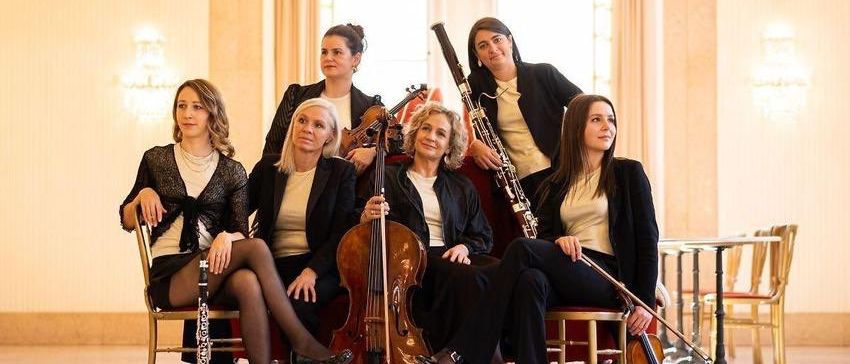 Eine Gruppe von Musikerinnen sitzt in einem Saal. Sie halten verschiedene Musikinstrumente, darunter Cello, Geige und Klarinette. Sie sind in formeller Kleidung gekleidet.