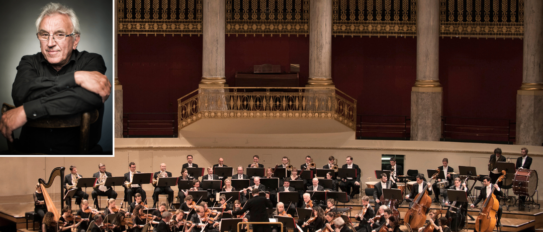 Ein Symphonieorchester tritt mit einem Dirigenten auf der Bühne auf. Musiker spielen Violinen, Violoncelli und Blechblasinstrumente. Der Veranstaltungsort hat kunstvolle Säulen und eine Galerie.