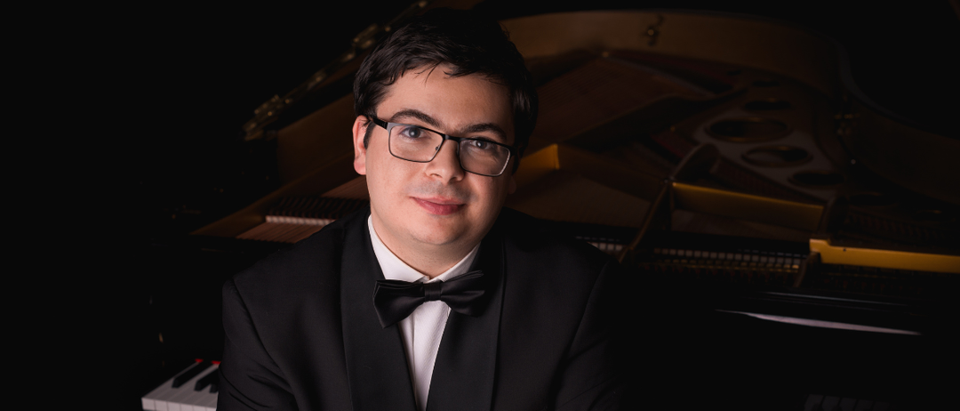Ein Mann in formaler Kleidung steht mit verschränkten Armen vor einem Flügel. Er trägt eine schwarze Fliege und eine Brille und lächelt leicht. Die Tasten des Klaviers sind im Vordergrund zu sehen.
