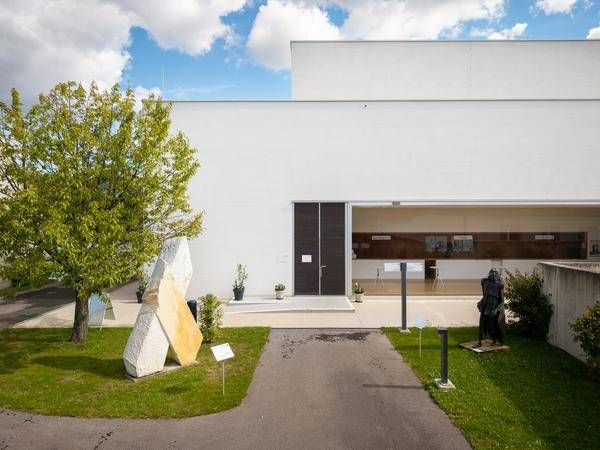 Ein weißes Gebäude mit einer großen offenen Tür und Fenstern steht neben einer Skulptur und Bäumen.