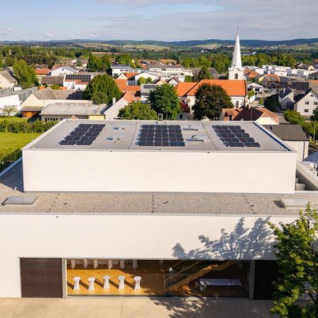 Luftaufnahme eines modernen Gebäudes mit Solarpanelen auf dem Dach, in einer malerischen Stadt gelegen, mit einer Kirche und Hügeln im Hintergrund.