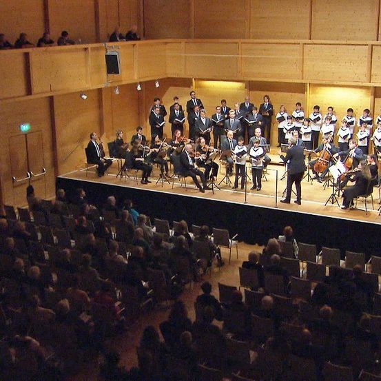 Ein Orchester und Chor spielen auf der Bühne in einem hölzernen Konzertsaal, mit einem Publikum darunter. Die Musiker spielen verschiedene Instrumente, und ein Dirigent dirigiert von vorne.