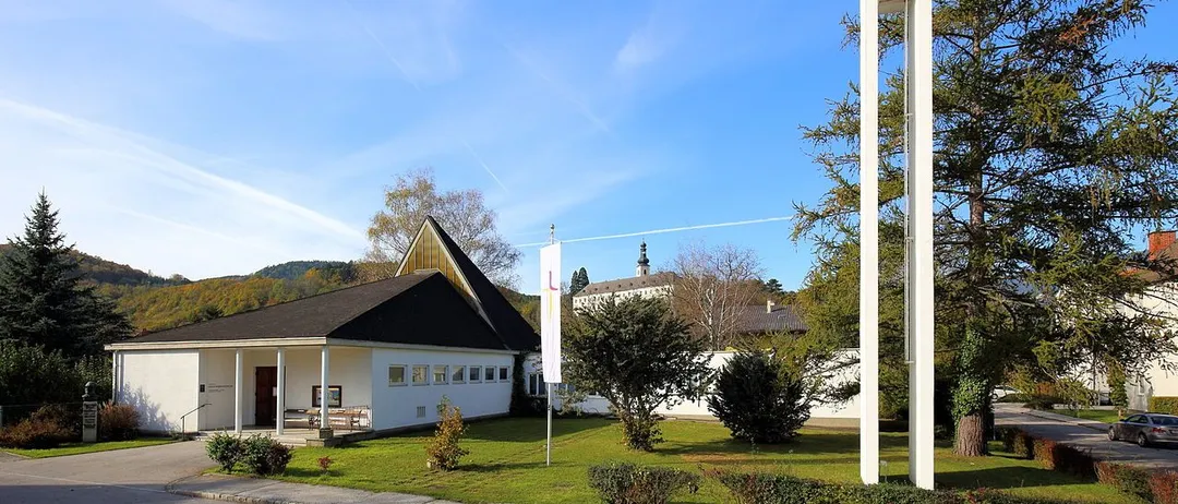 Eine moderne weiße Kirche mit einem Turm und einem Kreuz, umgeben von Grün und einem klaren blauen Himmel.