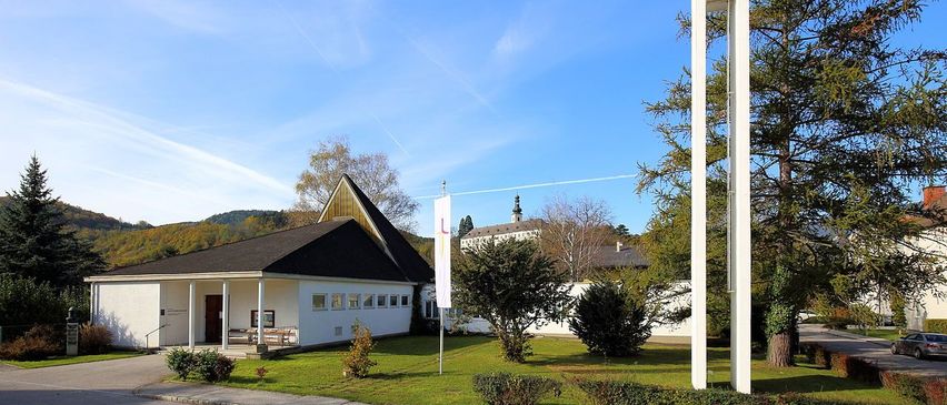 Eine moderne weiße Kirche mit einem Turm und einem Kreuz, umgeben von Grün und einem klaren blauen Himmel.