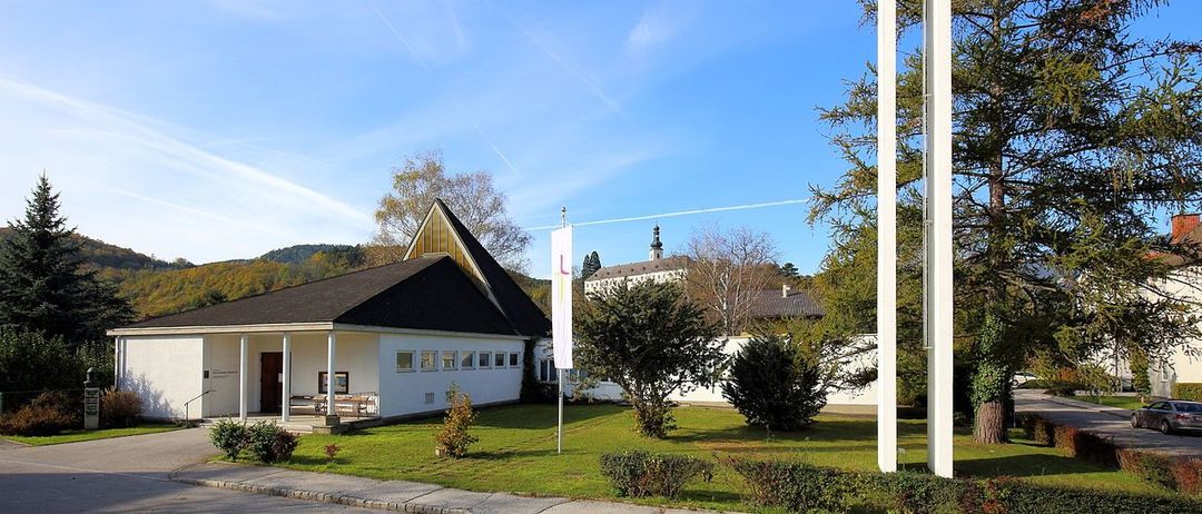 Eine moderne weisse Kirche mit einem Turm und einem Kreuz darauf, umgeben von Bäumen und Büschen.